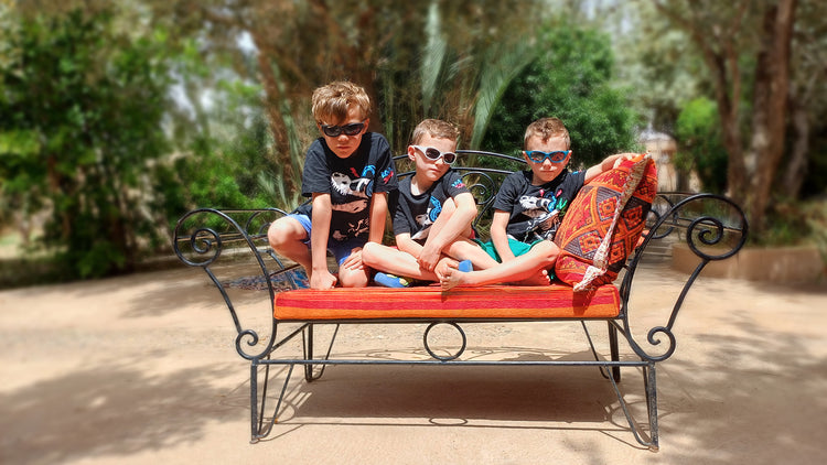 Drei Jungen mit ActiveSol Kids@school Sonnenbrille sitzen auf einer rot-orangenen Bank in einer Oase