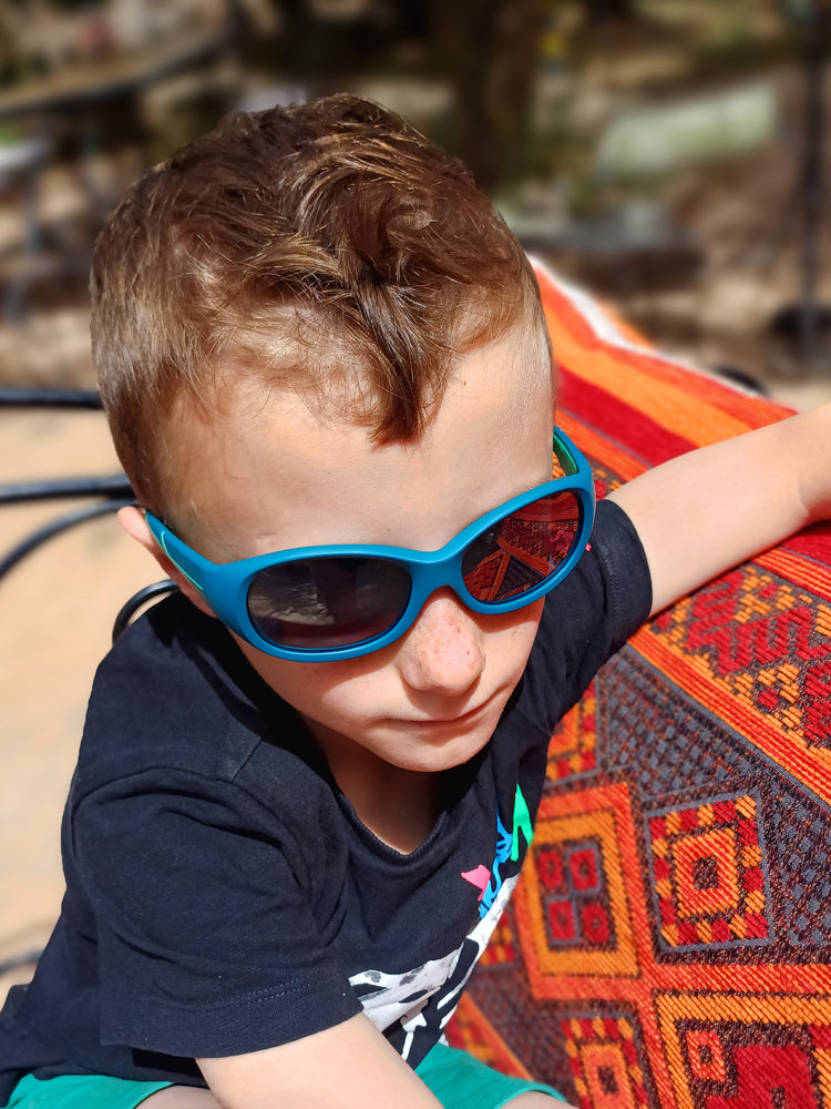 Ein Junge mit einer ActiveSol Kids@school Sonnenbrille sitz auf einer rot-orangenen Bank in einer Oase