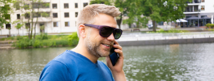 Lachender Mann mit Überzieh-Sonnenbrille. Er telefoniert am Wasser vor einer Promenade und nimmt gerne auch Dein Gespräch entgegen.